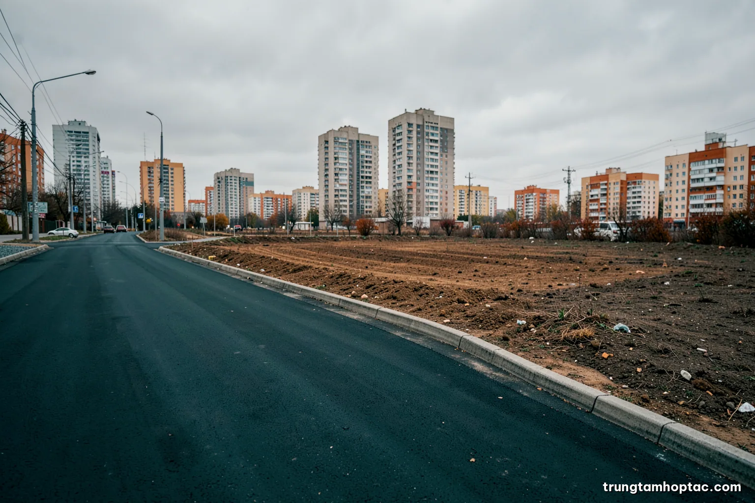 Low angle shot of a newly paved urban boulevard beside a vacant cleared land plot, distant apartment towers, overcast sky, desaturated grey and ochre tones