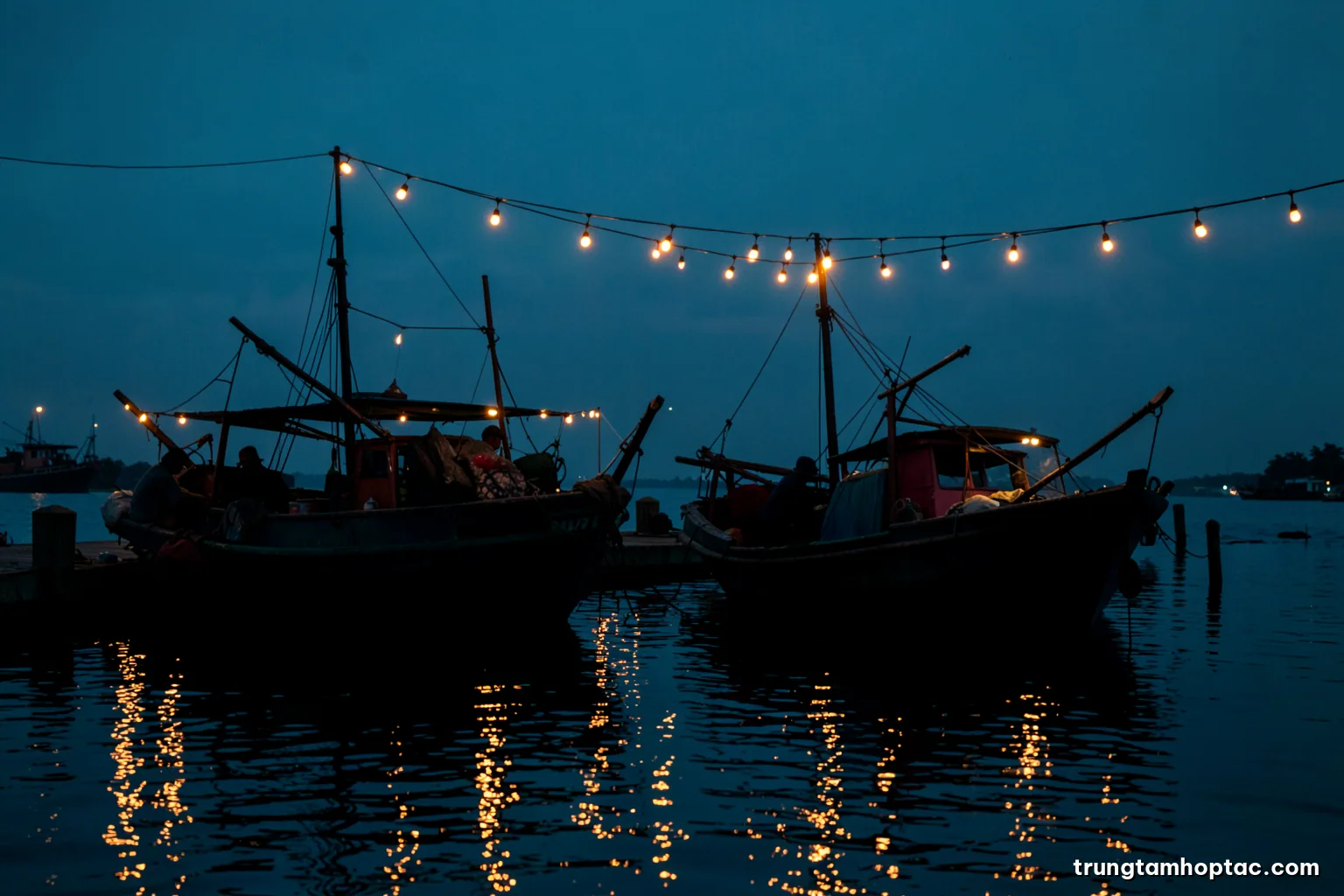 Silhouettes of two fishing boats side by side unloading catch at a shared pier in Ca Mau at blue hour, string lights reflecting on calm water, deep navy and amber palette