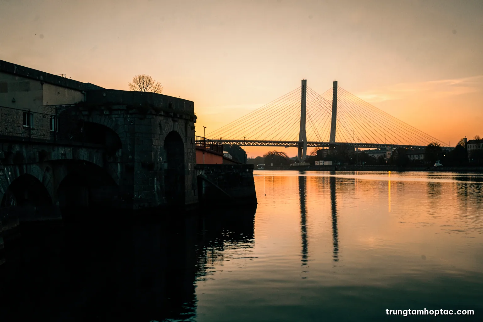 Silhouette of an old stone canal lock and arched bridge at sunset with a modern cable-stayed bridge on the horizon, calm water reflections, sepia and copper palette