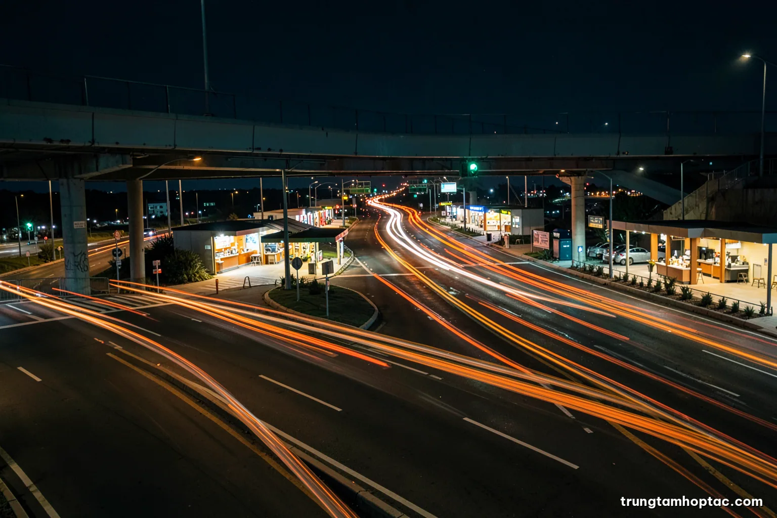 Night long exposure of light trails passing through an expressway toll plaza seen from an overpass, rows of booths glowing, deep navy and sodium orange palette