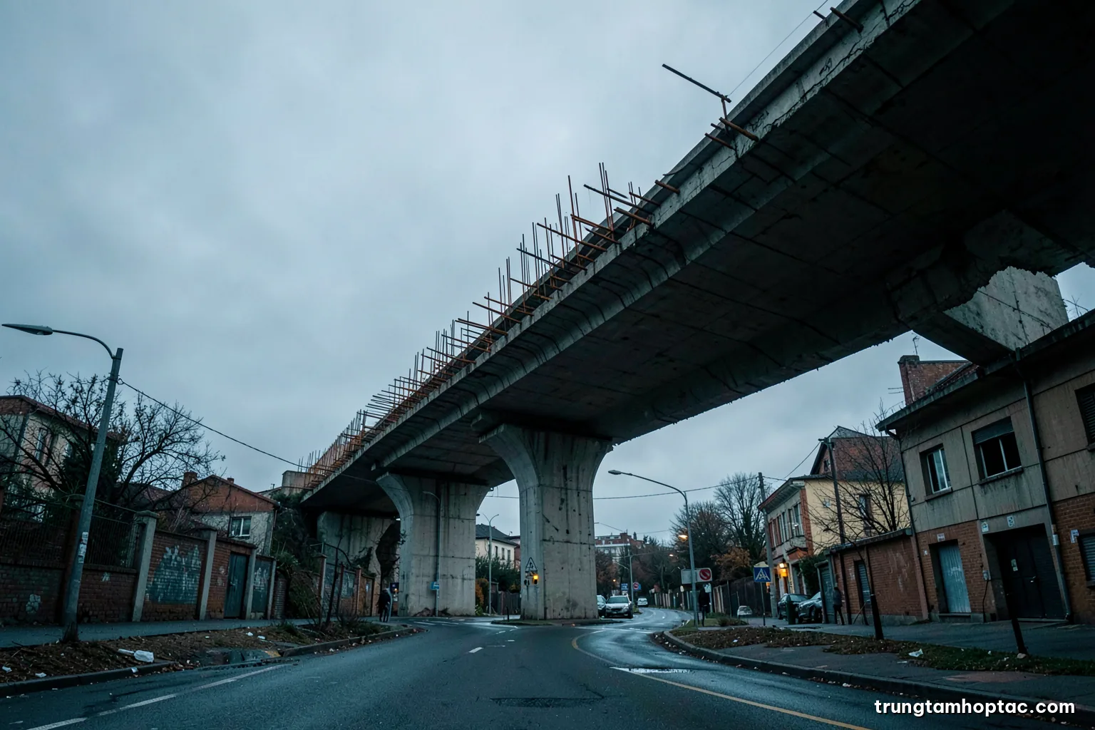 Low angle view of an unfinished elevated concrete viaduct with exposed rebar ending in mid-air above a quiet street, overcast dusk, cold grey and rust palette