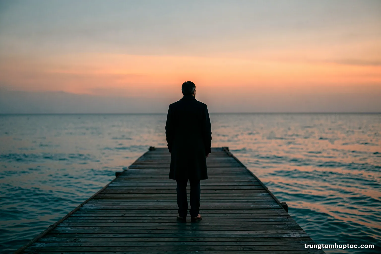 A lone figure in a dark coat standing at the end of an empty pier at dusk, shot from behind with a wide lens, calm grey sea and fading orange horizon